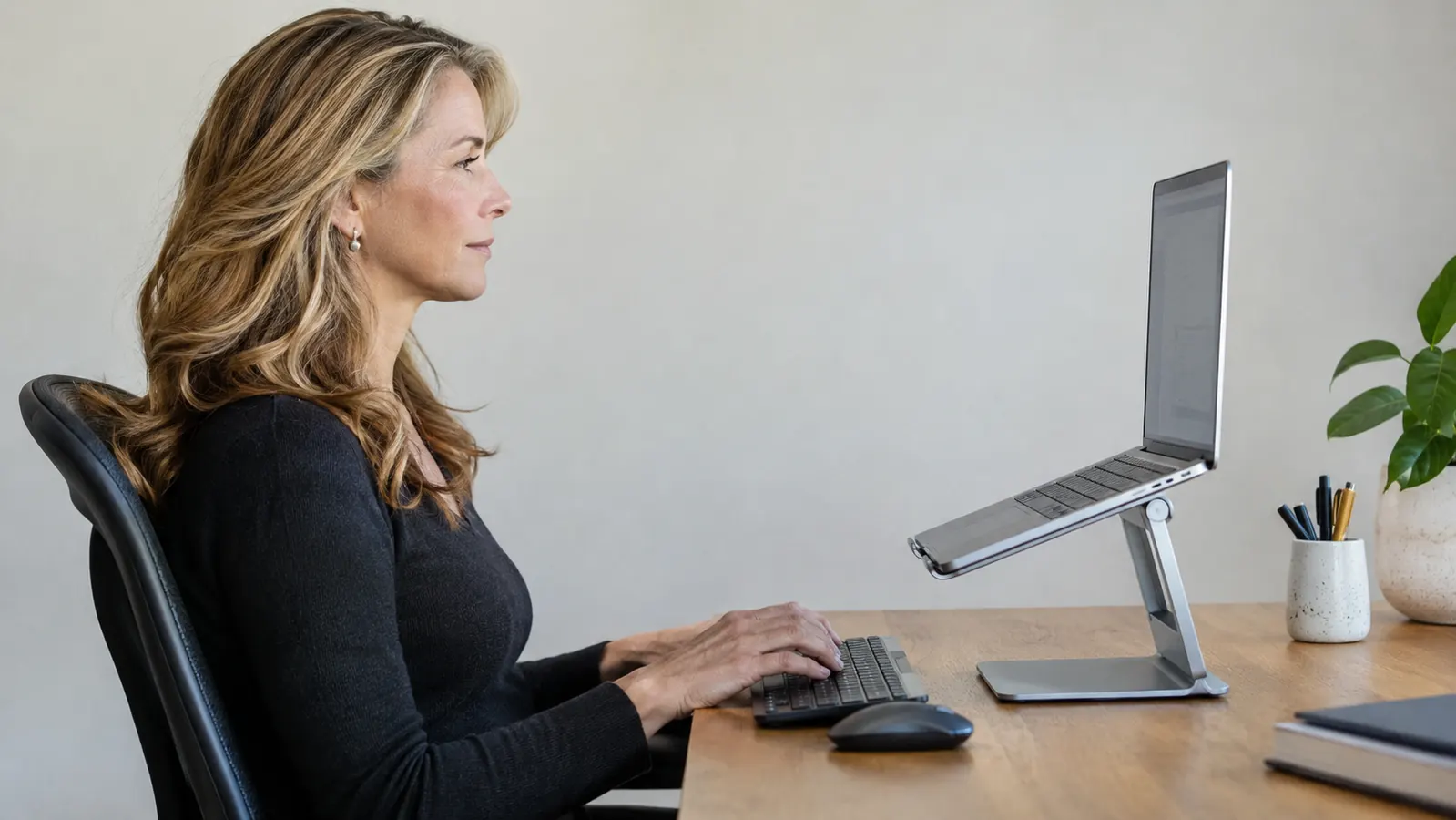 Side view of a woman in her fifties at a desk, laptop screen at eye level, head level and shoulders relaxed