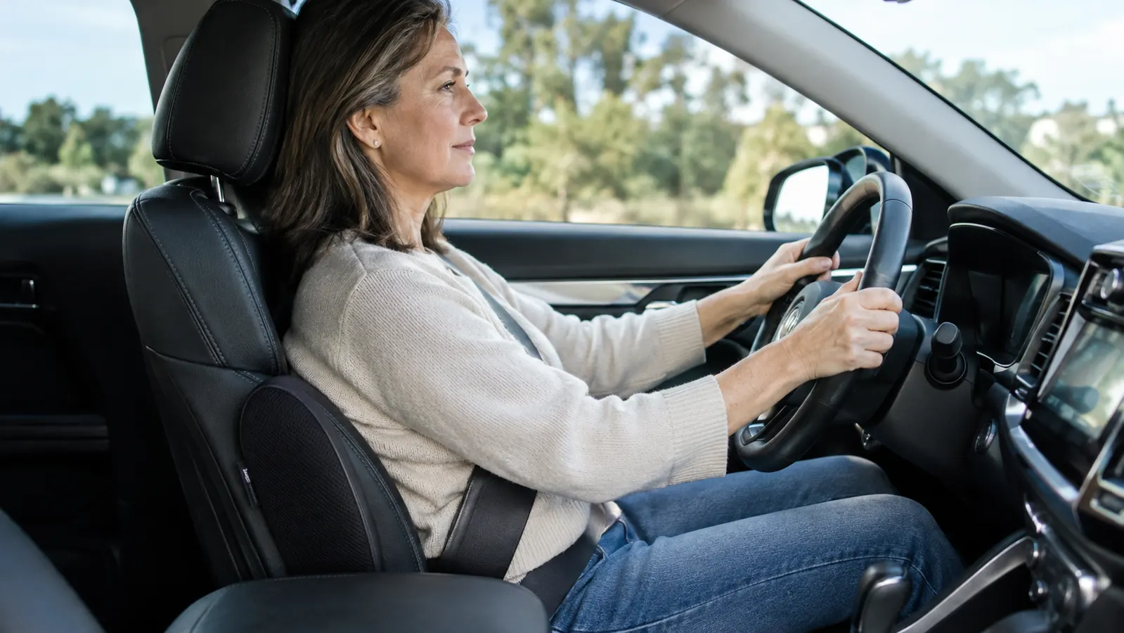 A woman in her fifties sitting upright in a car seat with a lumbar support pillow behind her lower back, hands relaxed on the wheel